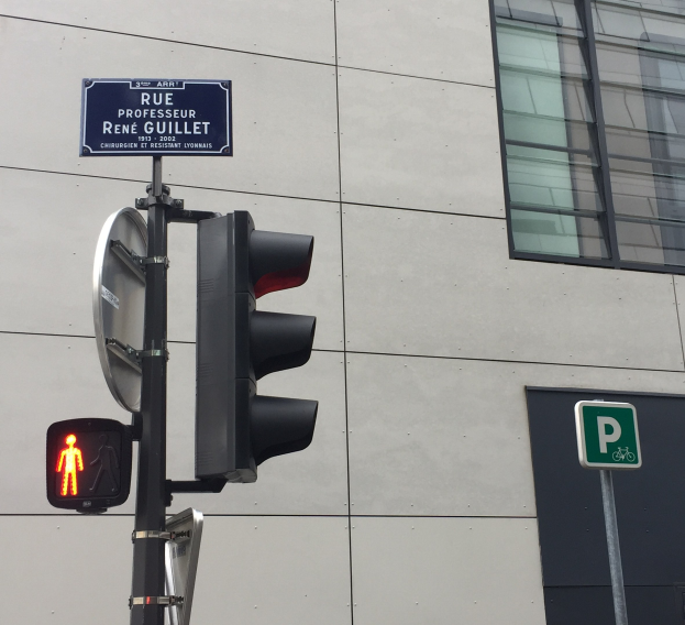 Rotes Verkehrslicht an einer Stange mit einem über die Stra├če führenden Fu├čgänger-Schild, vor einem Hintergrundgebäude.