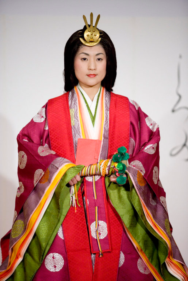 Eine Frau in einem traditionellen japanischen Kimono, das ein leuchtend rotes und grünes Kleid mit aufwändigen Mustern trägt, steht selbstbewusst auf einem Laufsteg mit ihrem Haar zu einem ordentlichen Knoten gebunden.