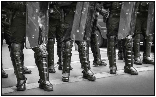 Schwarz-weißes Bild von Polizisten in einer Reihe, die Schilde halten, mit einem Fahrzeug im Hintergrund.