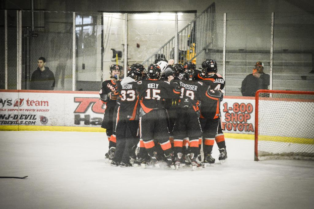 Eine Gruppe von Menschen in Eislaufschuhen, Handschuhen und Helmen steht auf einem Eisboden mit Absperrungen und Glastüren dahinter, einem Hockey-Tor mit Netz auf der rechten Seite und einem Hockey-Schläger auf der linken Seite.