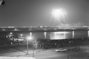 Ein Schwarz-Weiß-Foto einer Menge, die ein Feuerwerk über Wasser beobachtet, mit Fahrzeugen, Laternenmasten, Bäumen und Gebäuden im Hintergrund.