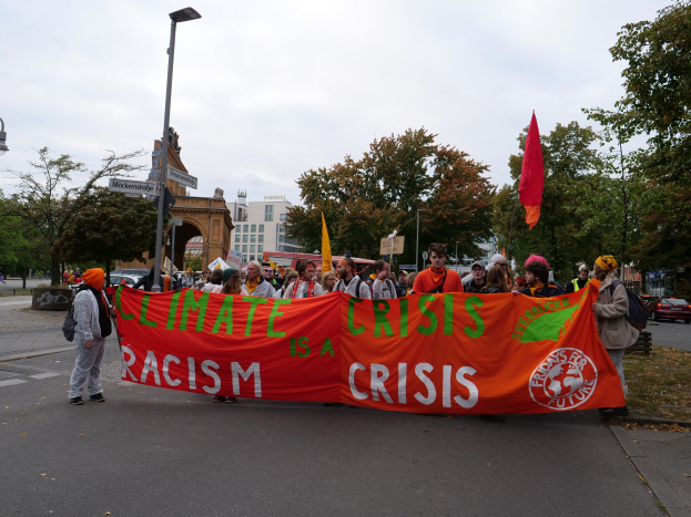 Eine Gruppe von Menschen marschiert eine baumbestandene Straße entlang und trägt ein Banner mit der Aufschrift "Klimakrise ist eine Krise", mit Fahrzeugen und Gebäuden im Hintergrund unter einem klaren blauen Himmel.