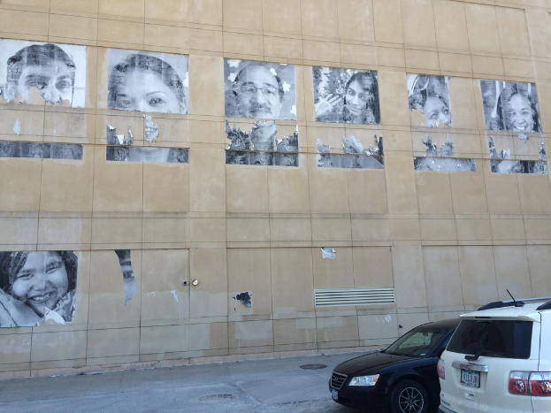 Ein Gebäude mit Autos auf der Straße davor, das Gullis und Plakate von Menschen an der Wand im Hintergrund zeigt.