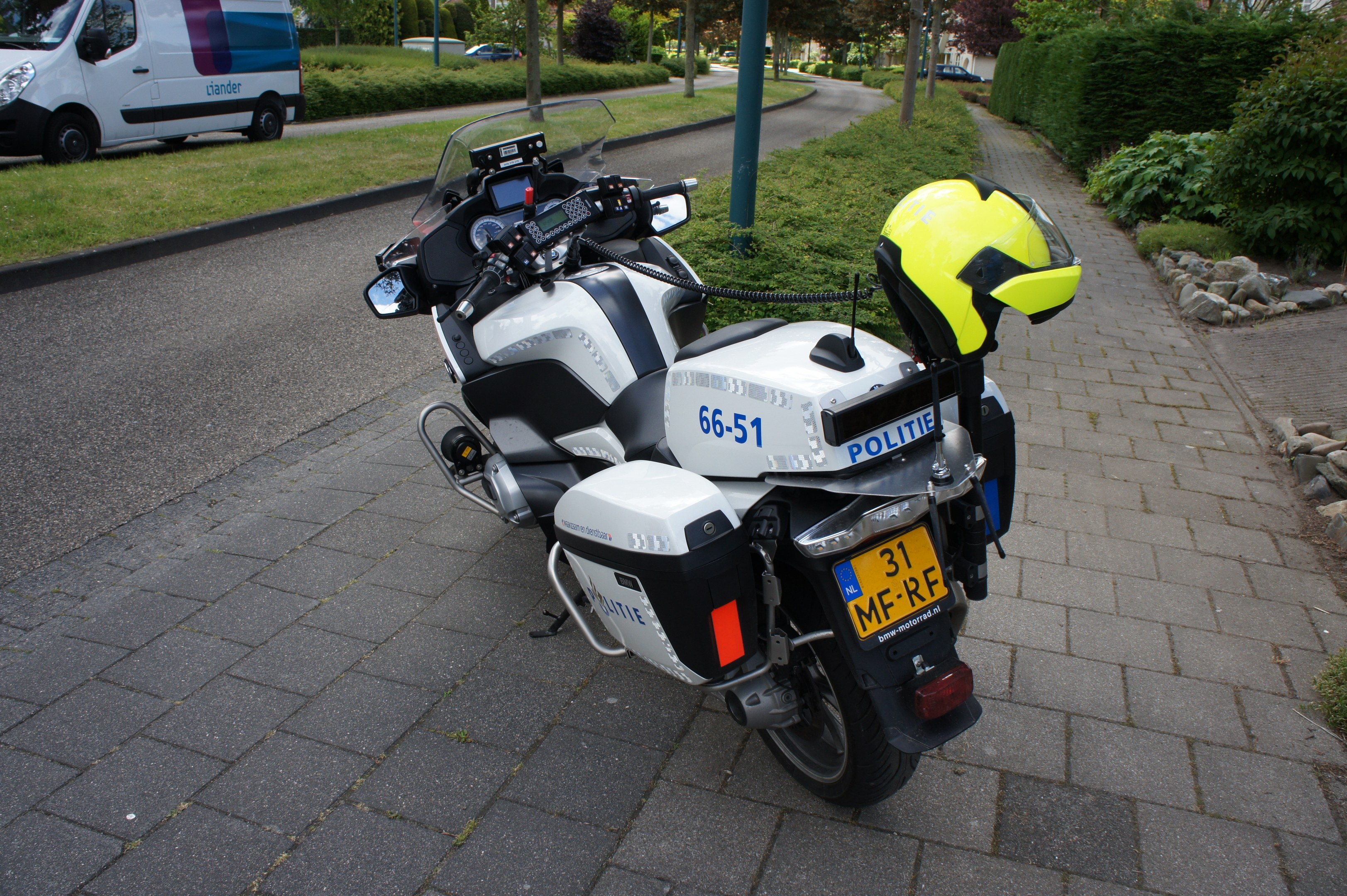 Ein auf der Straße geparktes Polizeimotorrad mit einem Helm darauf, umgeben von Bäumen, Pfählen, Gebäuden, Pflanzen, Gras, Steinen und anderen Fahrzeugen im Hintergrund.