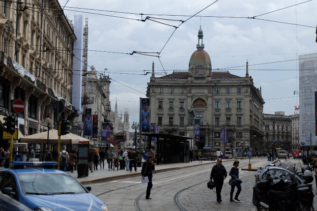 Eine belebte Straße mit einem parkenden Polizeiwagen, Passanten mit Taschen, fahrende Fahrzeuge, hohe Gebäude, Banner, Laternen und Ampeln unter einem bewölktem Himmel.
