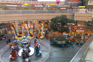 Ein Vergnügungspark innerhalb eines Einkaufszentrums mit verschiedenen Attraktionen, Bäumen, Menschen und Ständen, umgeben von Gebäuden oben und einem Wasserzeichen unten rechts.