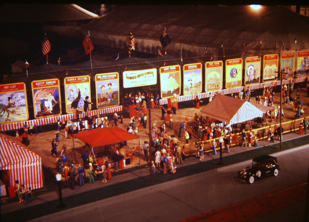 Ein altes Nachtfoto eines Karnevals mit Menschen, einem Auto, Polen mit Fahnen, einem Zaun, einem Zelt, Brettern mit Bildern und Text und einer Straßenlaterne.