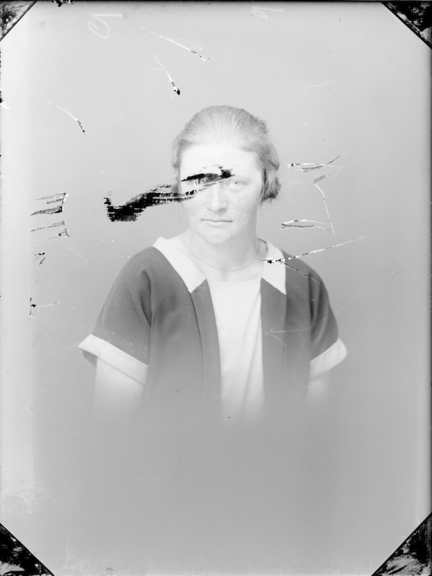 Ein Schwarz-Weiß-Foto einer Frau mit einem zerbrochenen Glasfenster dahinter, die direkt in die Kamera schaut und den Mund leicht öffnet.