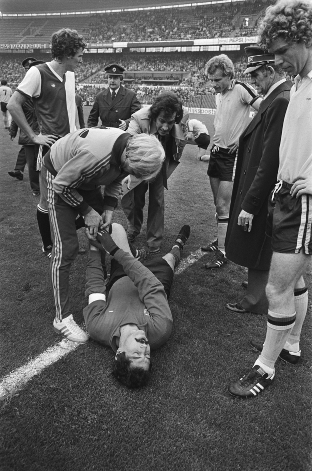 Schwarzes und wei├čes Bild eines Fussballspielers, der auf dem Boden liegt und von einer Gruppe M├Ąnner umgeben ist, mit einer Menge im Stadionhintergrund.