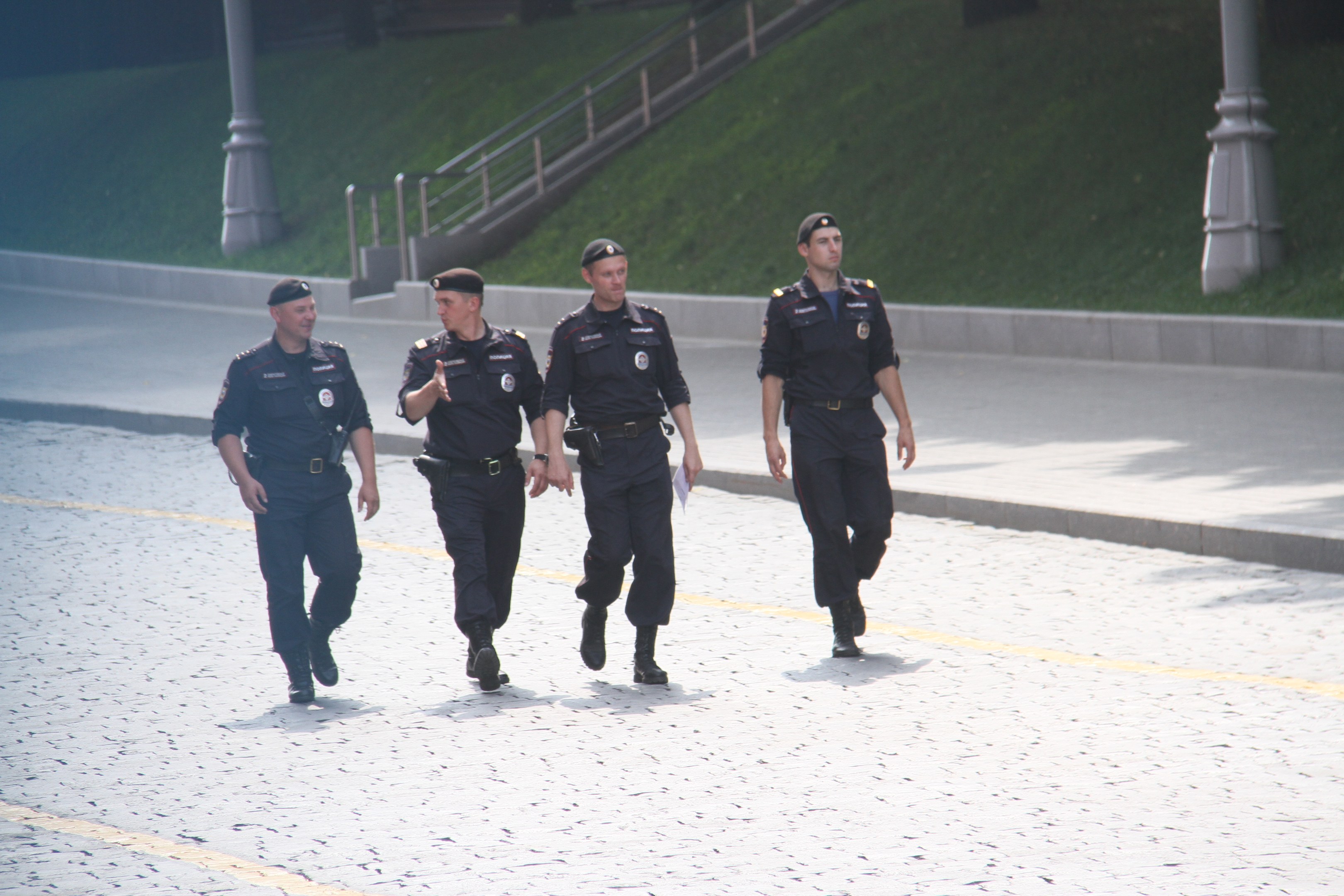 Drei Polizisten in Uniform und Mützen gehen eine Straße mit Geländern, Treppen und Gras im Hintergrund entlang.