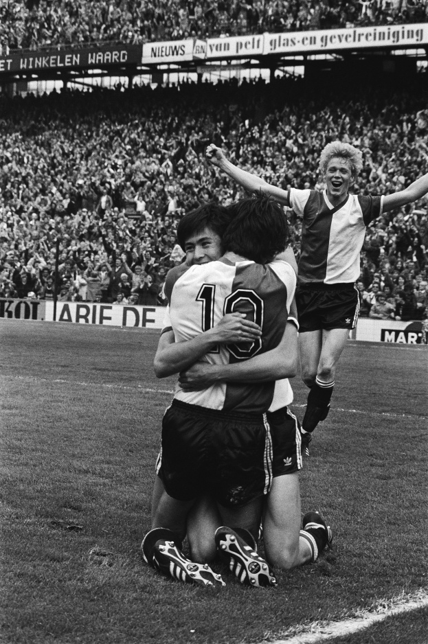 Drei Fussballspieler feiern ein Tor auf dem Boden vor einer jubelnden Menge in einer schwarz-weissen Stadionszene.