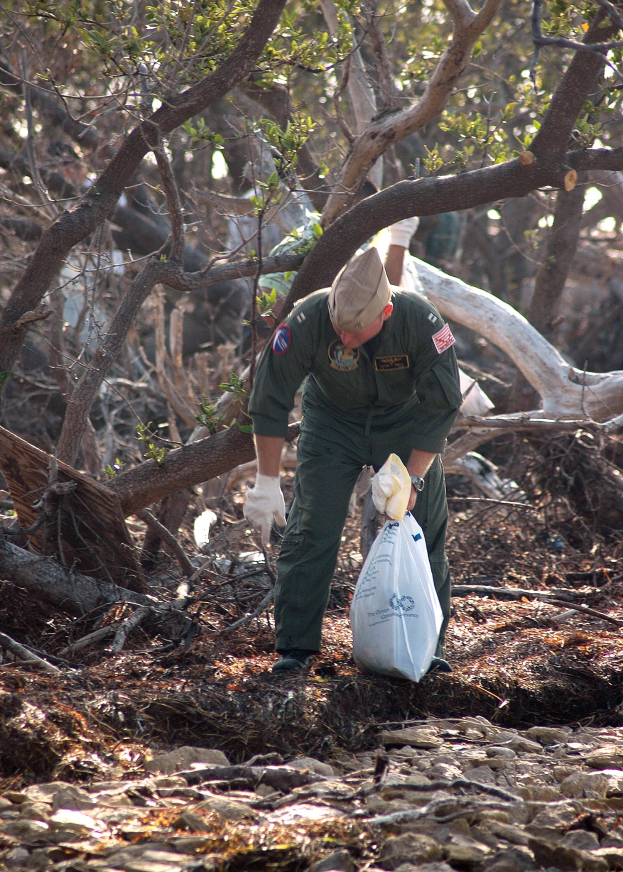 Ein Mann mit Mütze und Handschuhen h├Ąlt eine Polyethylenabdeckung, w├Ąhrend er Müll in einem wüldlichen Gebiet mit Steinen und trockenen Bl├Ąttern auf dem Boden sammelt.