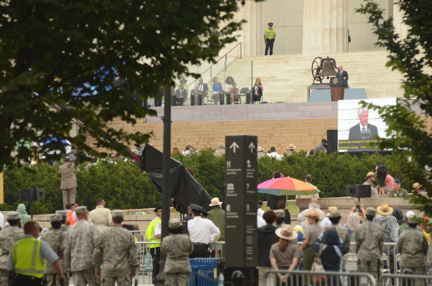 Army personnel and civilians stand and sit listening to a speech given by the president, with trees, bushes, and stairs in the background.
