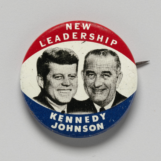 Ein Ansteckbutton auf einer weißen Oberfläche zeigt zwei Bilder von Kennedy und Johnson mit dem Text 'Neue Führung' darunter.