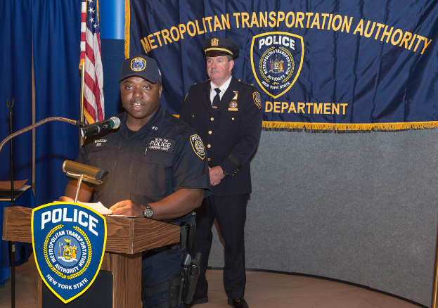 Zwei Polizeibeamte stehen an einem Rednerpult, einer hält Papiere und spricht in ein Mikrofon, mit einer Fahne, einem Banner und einem Tisch im Hintergrund.