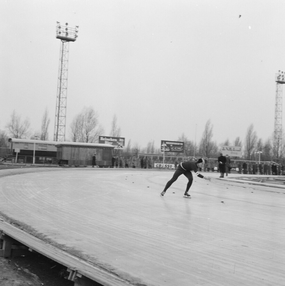 Schwarz-weiß-Foto eines Mannes, der auf einer Eisbahn mit Zuschauern, einer Hütte, einem Zaun, einer Fahne, Pfosten, Bäumen, Lichtern auf Türmen und einem bewölkten Himmel Schlittschuh läuft.