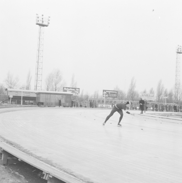 Schwarz-weiß-Foto eines Mannes, der auf einer Eisbahn mit Zuschauern, einer Hütte, einem Zaun, einer Fahne, Pfosten, Bäumen, Lichtern auf Türmen und einem bewölkten Himmel Schlittschuh läuft.