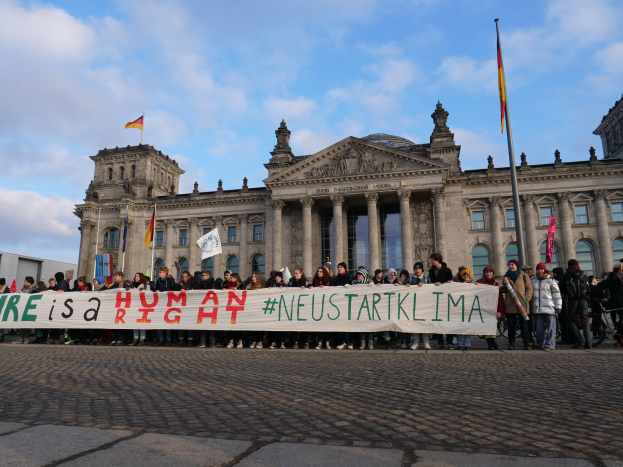 Eine Gruppe von Menschen steht vor dem Reichstaggebäude in Berlin, Deutschland, mit einer Fahne, auf der 'Wir sind ein Menschenrecht' steht, während das Gebäude mit Säulen, Fenstern, Bögen und Statuen verziert ist und von Fahnen an Stangen umgeben ist, unter einem bewölkten Himmel.