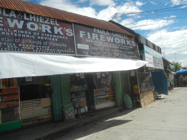 Ein Geschäft mit einer "Feuerwerk"-Schriftzug, umgeben von verschiedenen Gegenständen und Gebäuden, mit Bäumen, Pfählen, Drähten und einem bewölkten Himmel im Hintergrund.