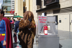 Eine Menschenmenge, darunter einige in Kostümen, versammelte sich vor einem Gebäude mit sichtbarer Beschilderung und Türen, mit einer Kiste und einem Feuerlöscher in der Nähe.