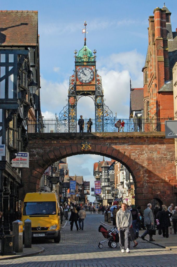 Eine belebte Straße mit Fußgängern, ein gelber Van auf dem linken Gehweg, eine Brücke mit zwei Personen darüber, eine Uhr über der Brücke, Gebäude auf beiden Seiten und der Himmel darüber.
