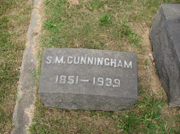 Ein Grabstein mit Text darauf auf einer grasbewachsenen Fläche, mit einem anderen Stein rechts daneben.