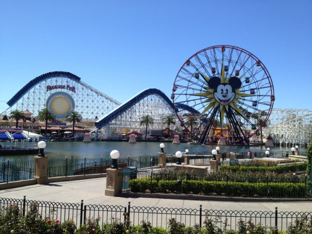 Disneylands California Adventure Vergnügungspark mit einem Riesenrad, Achterbahn, Grünflächen, Laternenmasten, einem Gewässer und einem hellblauen Himmel.