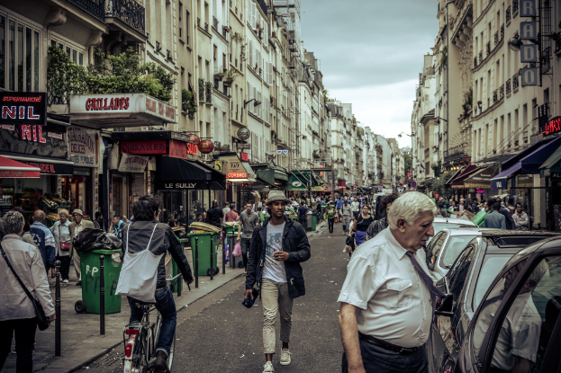 Zwei große Gebäude stehen sich gegenüber auf einer belebten Straße mit Fußgängern, Händlern und parkenden Autos, während ein Mann ein Fahrrad fährt und Mülltonnen entlang des Bürgersteigs aufgestellt sind.