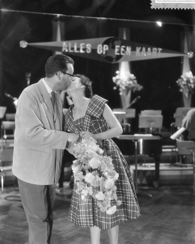 Ein Mann und eine Frau umarmen und küssen sich auf einer Bühne, wobei die Frau einen Blumenstrauß hält, umgeben von Stühlen und Blumenvasen.