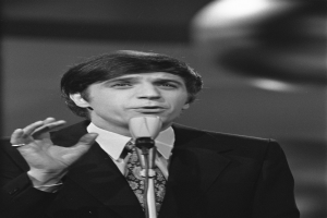 Ein Mann in einem schwarzen Anzug, weißem Hemd und gemustertem Schlips spricht in ein Mikrofon, mit ernstem Gesichtsausdruck und zurückgekämmtem Haar, vor einem unscharfen Hintergrund.