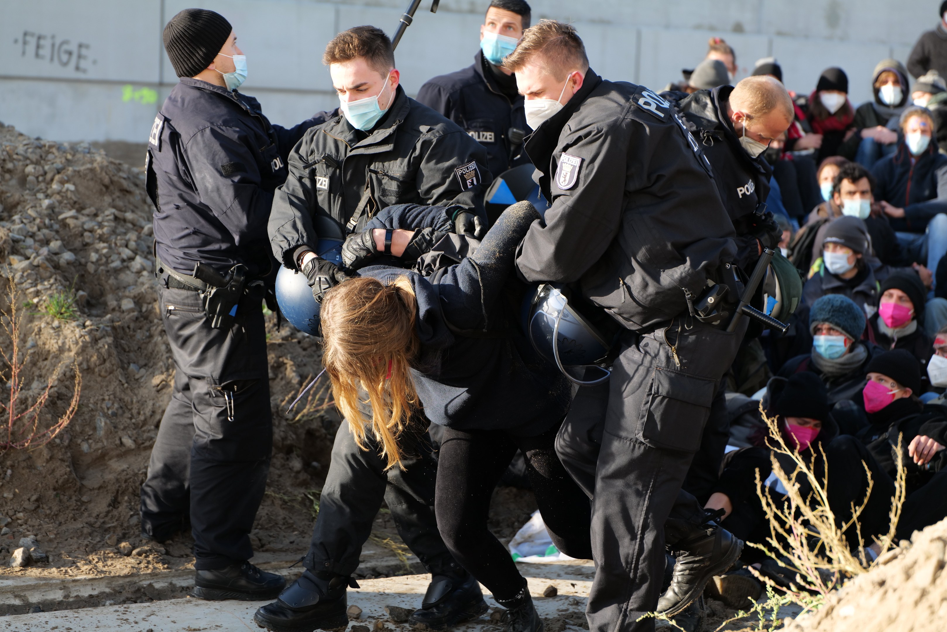 Gruppe maskierter Personen in Mützen, einige bewaffnet, die eine Frau umgeben, die von der Polizei begleitet wird, mit städtischer Vegetation und einer protestbezogenen Wandnachricht im Hintergrund.