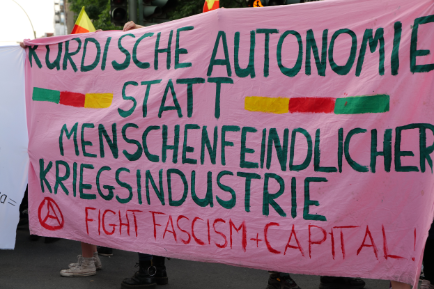 Eine Gruppe von Menschen steht auf einer Straße und hält ein pinkes Banner mit der Aufschrift "Kurdische Autonomie Statt Menschenfeindlicher Kriegsindustrie Fight Fascism Capital", mit Ampeln, Flaggen, Bäumen, Gebäuden und einem klaren blauen Himmel im Hintergrund.