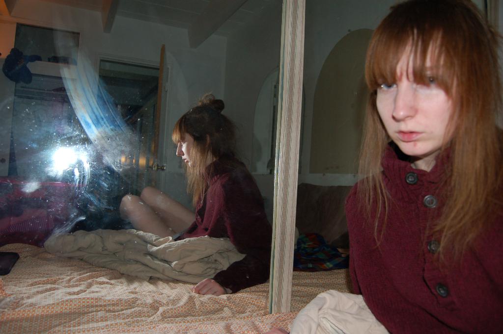 Eine Frau in roter Jacke steht neben einem Spiegel, der ihr Bild reflektiert, mit einem Bett und seiner Bettwäsche im Hintergrund.