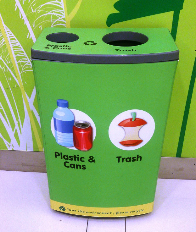 Ein grüner Mülltonne aus Plastik und Dosen auf einem weißen Boden, mit einem Plakat an der Wand im Hintergrund.