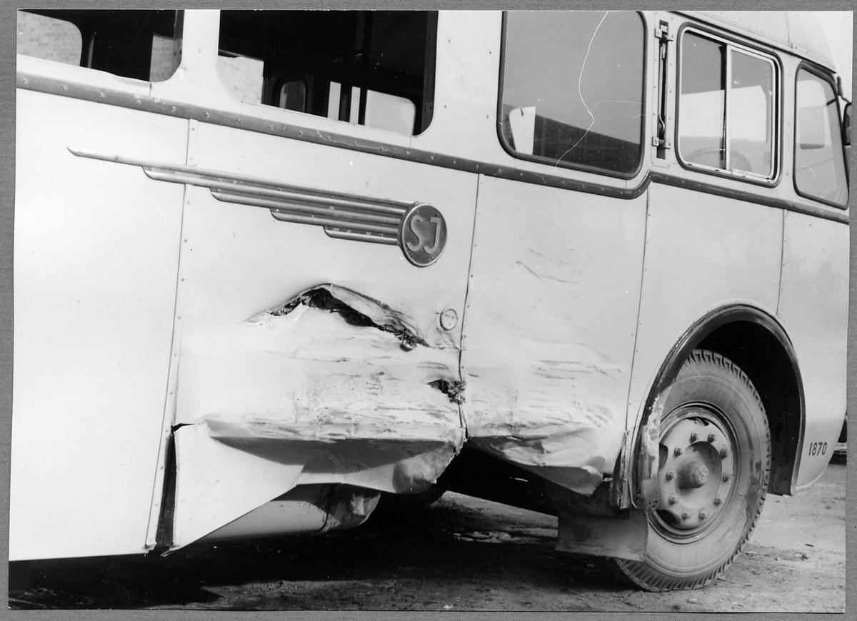 Schwarzes und weißes Foto eines beschädigten Busses am Straßenrand mit eingedrückter Front, zersplitterten Fenstern und einer offenen Fahrertür, die den Innenraum zeigt.