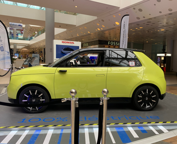 Ein gelbes wasserstoffbetriebenes Elektroauto steht drinnen vor einem Gebäude, umgeben von Absperrpollern, Bannern und Schildern, mit einer Person darin und einer Gruppe von Menschen in der Nähe.