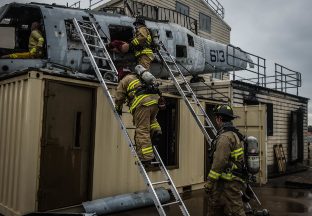 Feuerwehrleute in Uniform und Helmen arbeiten an einem Hubschrauber auf einem Gebäudedach, mit Leitern, einem Sauerstoffzylinder, einem Container, Bäumen und einem klaren Himmel im Hintergrund.