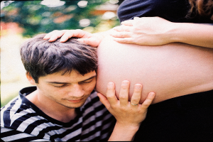 Ein Mann und eine Frau stehen nebeneinander, der Mann berührt sanft den Bauch der Frau, der Hintergrund ist unscharf.