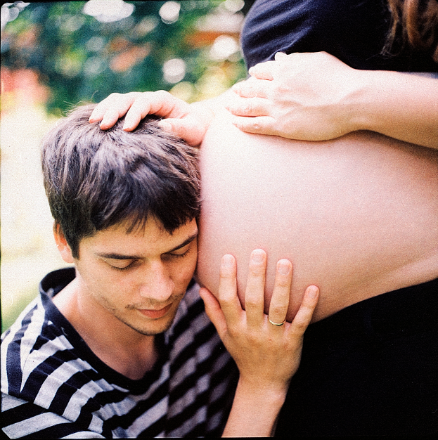 Ein Mann und eine Frau stehen nebeneinander, der Mann berührt sanft den Bauch der Frau, der Hintergrund ist unscharf.