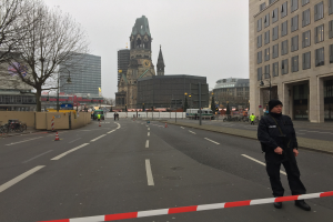 Polizist in schwarzer Uniform und Mütze auf einer Straße in Berlin mit Verkehrskegeln, Fahrrädern, Laternenpfählen, Bäumen, Gebäuden, einem Kirchturm und einem klaren blauen Himmel im Hintergrund.