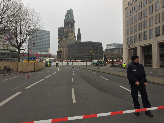 Polizist in schwarzer Uniform und Mütze auf einer Straße in Berlin mit Verkehrskegeln, Fahrrädern, Laternenpfählen, Bäumen, Gebäuden, einem Kirchturm und einem klaren blauen Himmel im Hintergrund.