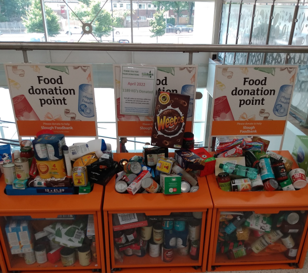 Orange Schrank Essensspende-Station mit Regalen voller Flaschen, Dosen, Schachteln und anderen Gegenständen, mit informativen Schildern, durch Glasfenster mit Außenfahrzeugen, Bäumen, Gebäuden und Himmel.