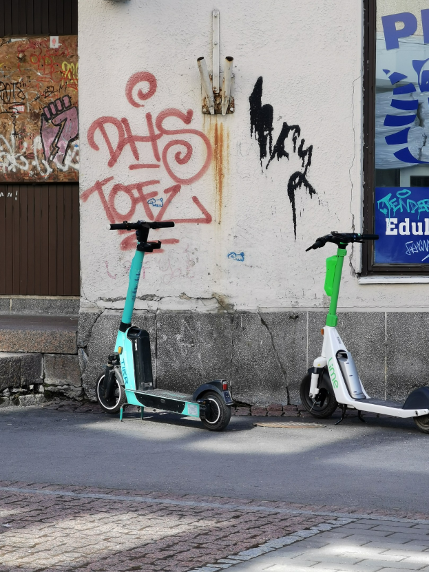 Zwei Elektro-Scooter nebeneinander auf einer Straße geparkt, mit einer Graffiti-bedeckten Wand und einem Plakat im Hintergrund.
