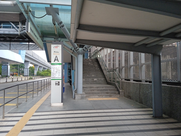 Bahnsteig mit Treppe, Straße mit Geländer auf der linken Seite, Hintergrund mit Bäumen, Pfählen, Gebäuden und einer Brücke, mit Rohren und einer Tafel oben auf dem Bahnsteig.