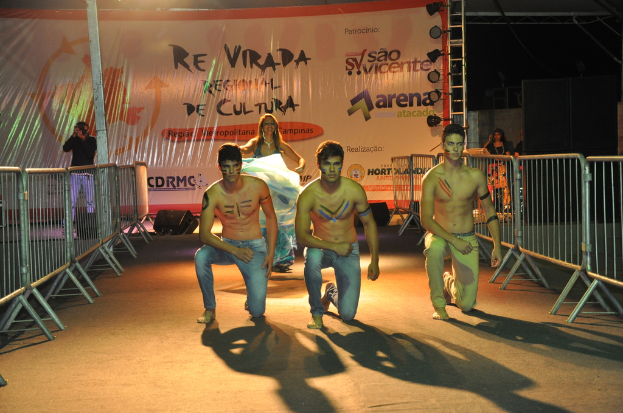 Drei Jungen ohne Hemd knien auf dem Boden während eines Kulturereignisses, mit Grills auf beiden Seiten und einem großen Banner im Hintergrund, während eine Person links unter Scheinwerfern ein Foto macht.