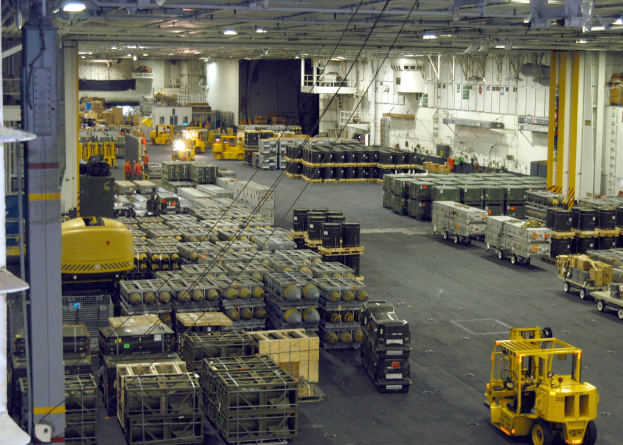 Ein großes Lagerhaus voller Numerous Trucks, Container, Menschen, Lichter, Stangen und andere Gegenstände mit einem prominenten Rad.