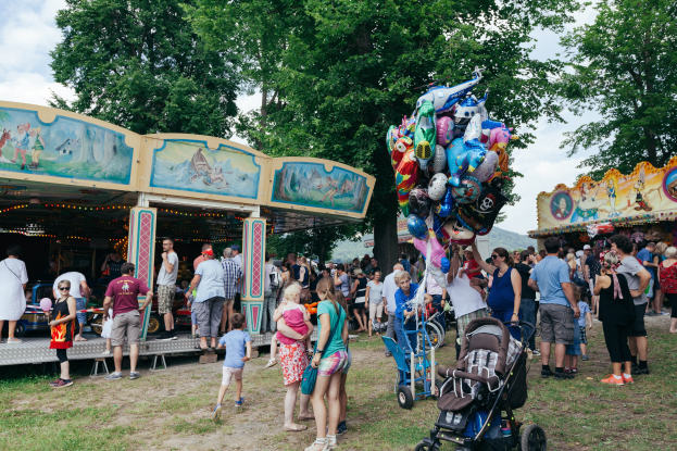 Eine Menschenmenge um einen bunt beleuchteten Karussellschlitten auf einem Volksfest, einige mit Kinderwagen, mit Bäumen und einem bewölktem Himmel im Hintergrund.