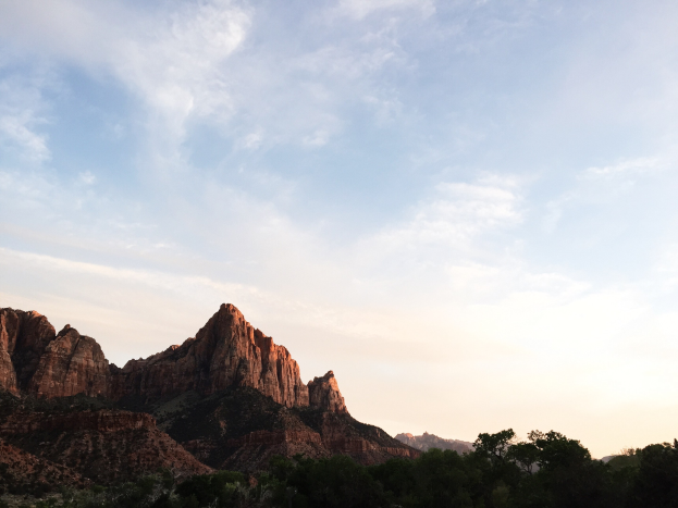 Ein malerischer Blick auf den Zion-Nationalpark in Utah mit Bäumen, Hügeln und einem Himmel mit Wolken, beleuchtet von der untergehenden Sonne.
