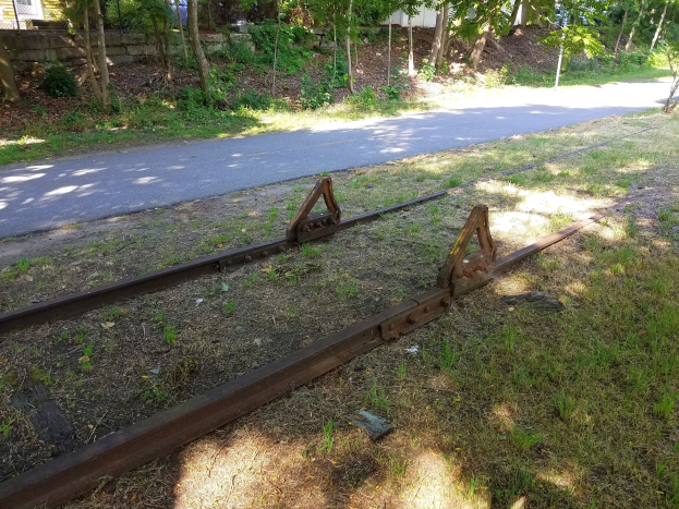 Eisenbahnschiene an einer Straße in Reparatur, umgeben von Gras und Bäumen, mit einer Wand und einem Gebäude im Hintergrund.