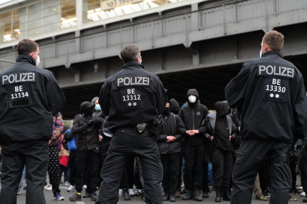 Polizeibeamte in schwarzer Uniform und Masken stehen vor einer Menge während einer Demonstration, mit einer Brücke und einem Gebäude im Hintergrund.